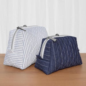 Op de afbeelding: Twee gewatteerde etuis met ritssluiting. De grotere etui heeft een lichtblauw en wit gestreept patroon, terwijl de kleinere etui donkerblauw is met dunne witte verticale strepen. Beide items hebben zilverkleurige metalen ritsen en stiksels.