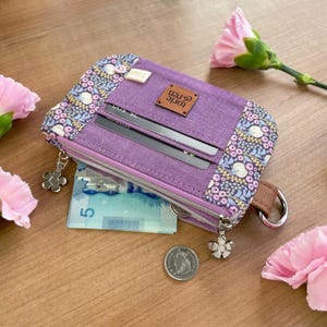 May include: A purple wallet with a floral pattern on the edges and a brown leather tag that reads "torie eren". The wallet has a zipper closure, a flower-shaped charm, and a brown wrist strap. Inside, there are credit card slots and a bill.