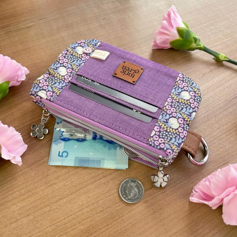May include: A purple wallet with a floral pattern on the edges and a brown leather tag that reads "torie eren". The wallet has a zipper closure, a flower-shaped charm, and a brown wrist strap. Inside, there are credit card slots and a bill.