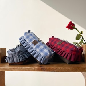 May include: Three fabric pencil cases with ruffled edges rest on a wooden surface. One is blue and white gingham, another is red plaid, and the third is blue and white striped. Each has a zipper and a small label that reads "little gurch".