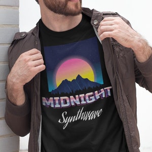 May include: Black t-shirt featuring a retro sunset design with mountains and the text "MIDNIGHT Synthwave". The graphic has a vibrant color palette of pink, yellow, and blue. The shirt is worn by a person.