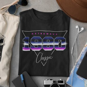 Puede incluir: Camiseta negra con un diseño retro wave que presenta el año 1980 en una fuente futurista y colorida. El texto "Retro Wave" y "Classic" también están incluidos en el diseño.