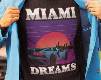 Könnte beinhalten: Schwarzes T-Shirt mit den weißen Worten "MIAMI DREAMS", das ein Retro-Design eines Autos, eines Sonnenuntergangs und einer Skyline zeigt. Die Person trägt einen hellblauen Blazer und eine Sonnenbrille.