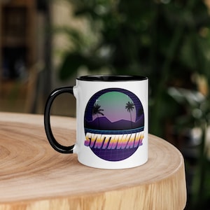 Può includere: Tazza in ceramica bianca con bordo e manico neri. La tazza presenta una grafica in stile retrò di un tramonto con palme e il testo "Synthwave" in un font colorato e pixelato.
