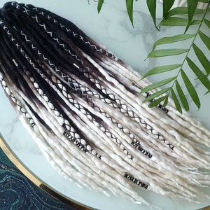 Puede incluir: Un conjunto de rastas sintéticas ombre negro y blanco con costuras y cuentas negras. Las rastas están dispuestas sobre una superficie blanca con un fondo verde y dorado.
