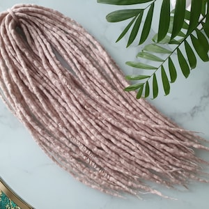 May include: A collection of light pink dreadlocks, displayed on a marble surface. The dreadlocks are thick and textured, with a slightly irregular appearance. Green leaves are visible in the background, adding a natural element to the composition.