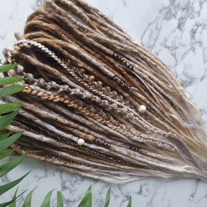 Puede incluir: Un conjunto de rastas sintéticas marrones, rubias y blancas con cuentas de madera. Las rastas están dispuestas en forma de abanico y están listas para ser instaladas.