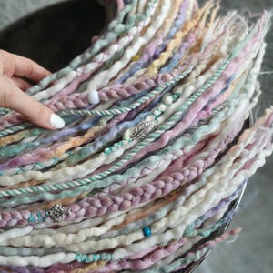 Könnte beinhalten: Eine Sammlung pastellfarbener Dreadlocks, mit Farbtönen von Rosa, Lavendel und Creme, mit Akzenten in Türkis und Pfirsich. Einige Dreadlocks sind mit Perlen und silbernen Charms verziert, was eine Boho-Ästhetik erzeugt. Die Dreadlocks sind kreisförmig angeordnet.