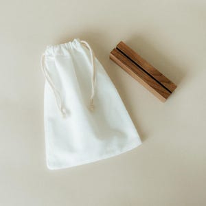 May include: A white drawstring bag and a wooden business card holder are displayed on a neutral background. The bag is made of a soft, opaque material. The card holder is rectangular with a dark stripe.