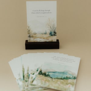 May include: A rectangular card featuring a watercolor landscape and the text I can do all things through Christ which strengtheneth me. PHILIPPIANS 4:13, displayed in a dark wooden stand, with a stack of similar cards featuring various landscapes.
