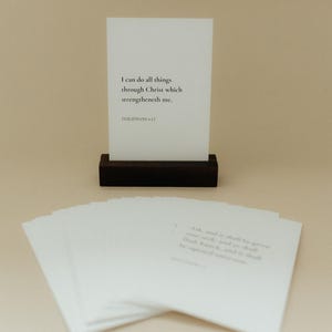 May include: A white rectangular card featuring the black printed text I can do all things through Christ which strengtheneth me. PHILIPPIANS 4:13, held upright in a dark brown wooden block stand, with a stack of additional white cards nearby.