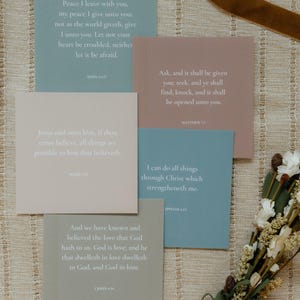 May include: Five square cards in muted tones of blue, pink, beige, and sage green, each featuring white printed Bible verses including John 14:27, Matthew 7:7, Mark 9:23, Philippians 4:13, and 1 John 4:16, arranged in a layered overlapping composition.