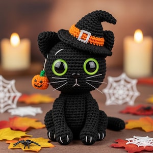 May include: A black crochet cat wearing a witch hat with an orange band and buckle. The cat has large green eyes, white whiskers, and a small pumpkin charm. The Halloween-themed cat is surrounded by fall leaves and spiderwebs.