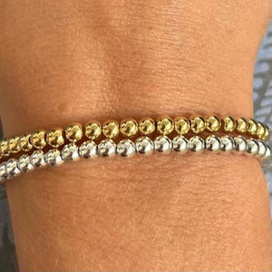 May include: Two beaded bracelets, one gold and one silver, stacked on a person's wrist.