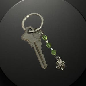 May include: A silver key with a key ring and a charm. The charm features a green beaded strand and a silver four-leaf clover. The key and charm are set against a dark background.