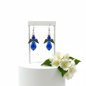 May include: A pair of blue angel earrings with silver-tone wings and accents. The earrings are displayed on a white stand, with white flowers and green leaves in the background. The earrings have a teardrop-shaped blue crystal.