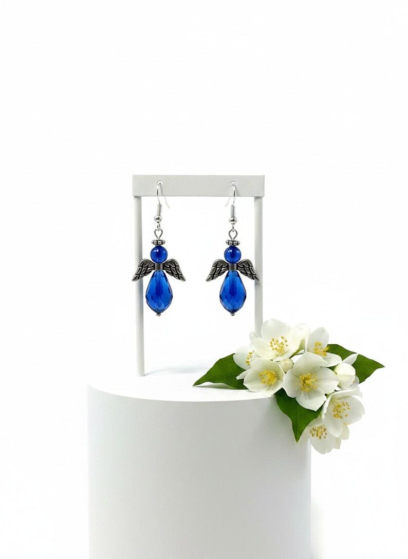 May include: A pair of blue angel earrings with silver-tone wings and accents. The earrings are displayed on a white stand, with white flowers and green leaves in the background. The earrings have a teardrop-shaped blue crystal.