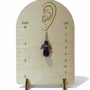 May include: A single dangle earring featuring a purple angel pendant with silver-colored wings and a faceted teardrop-shaped bead. The earring is displayed on a wooden stand with inch and centimeter measurements. The earring is approximately 1 inch (2.5 cm) in length.