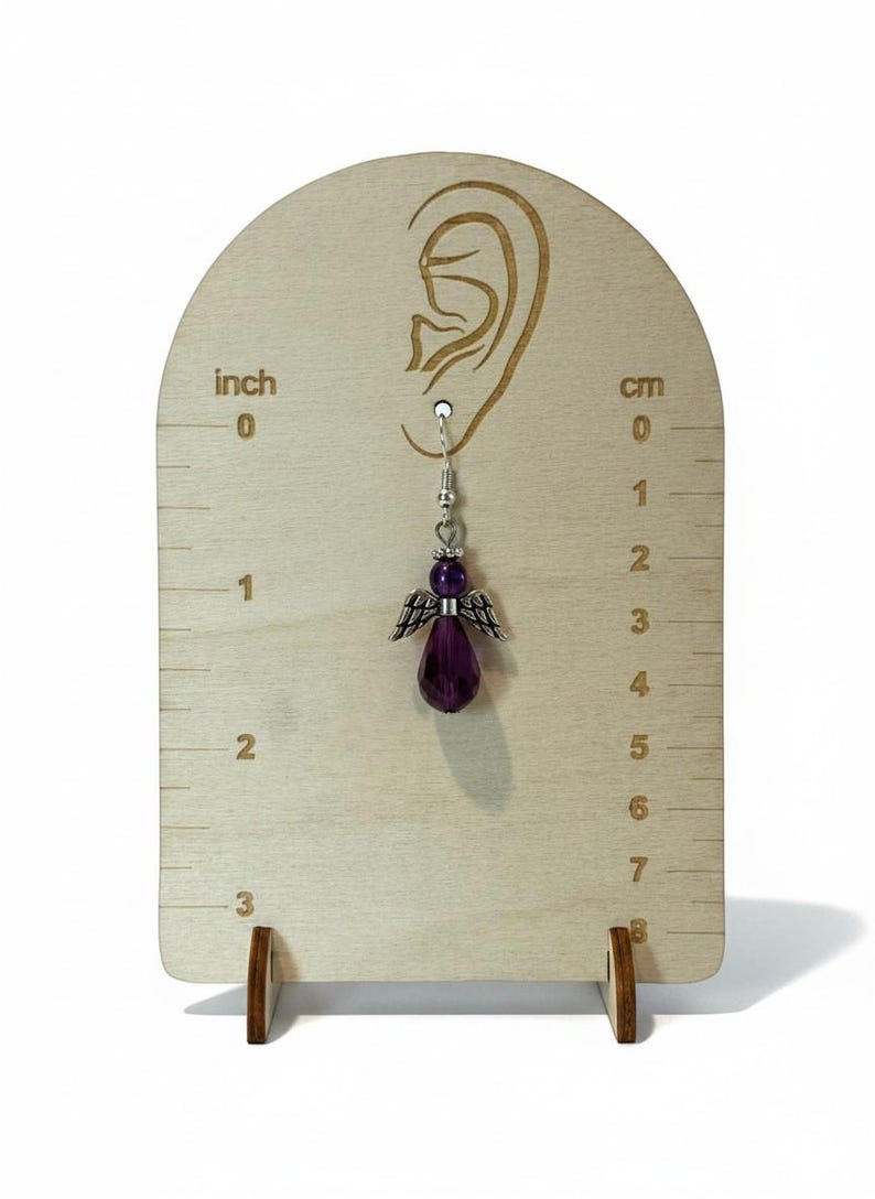May include: A single dangle earring featuring a purple angel pendant with silver-colored wings and a faceted teardrop-shaped bead. The earring is displayed on a wooden stand with inch and centimeter measurements. The earring is approximately 1 inch (2.5 cm) in length.
