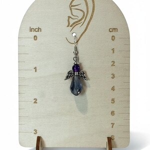 May include: A silver-toned angel-shaped earring with a faceted, gray-blue crystal pendant. The earring features a purple bead and silver wings. The earring is displayed on a wooden earring display stand with inch and cm measurements.