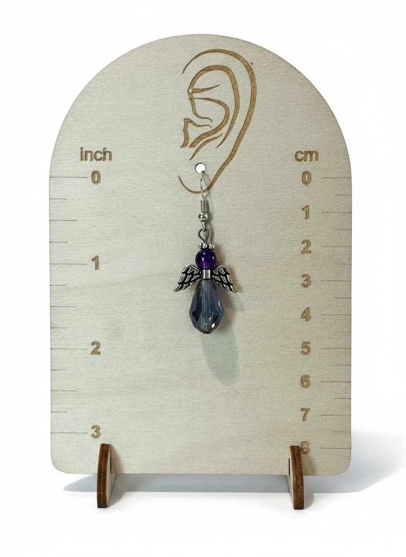 May include: A silver-toned angel-shaped earring with a faceted, gray-blue crystal pendant. The earring features a purple bead and silver wings. The earring is displayed on a wooden earring display stand with inch and cm measurements.