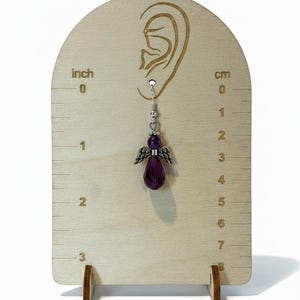 May include: A single angel-shaped earring with a purple gemstone body and silver wings is displayed on a wooden earring display. The display has inch and centimeter markings. The earring is approximately 1 inch in length.