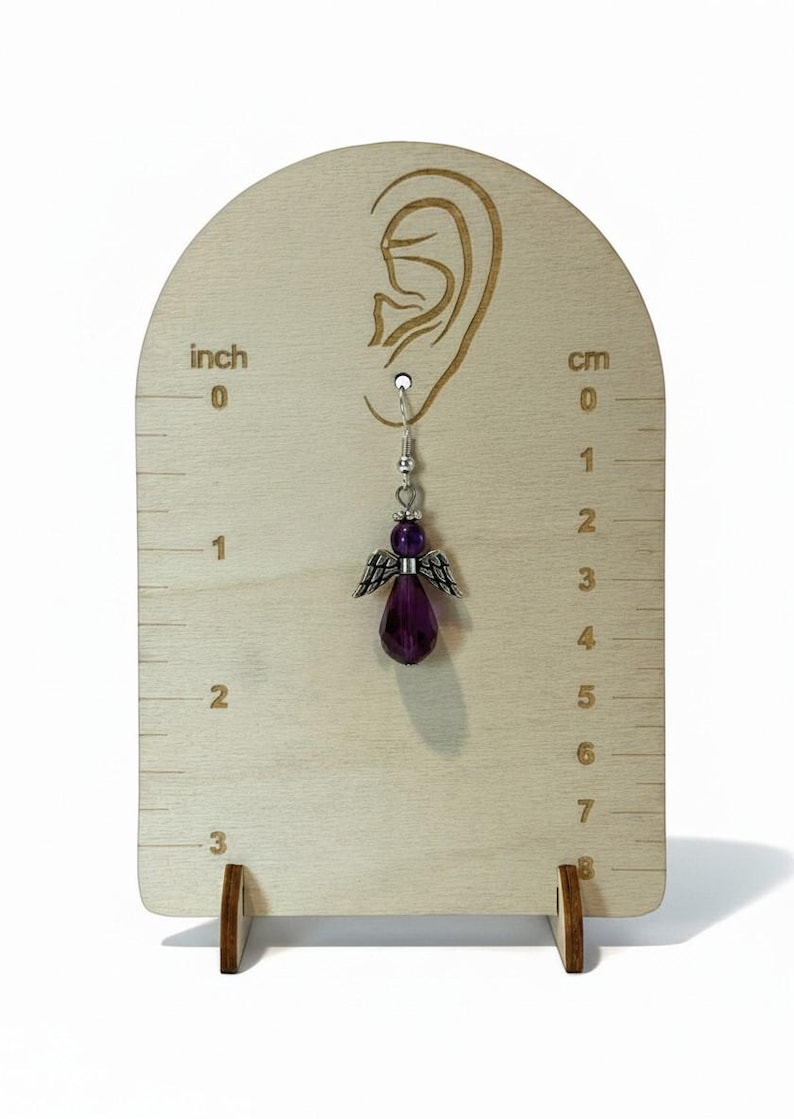 May include: A single angel-shaped earring with a purple gemstone body and silver wings is displayed on a wooden earring display. The display has inch and centimeter markings. The earring is approximately 1 inch in length.
