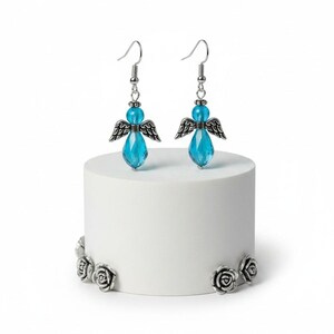 May include: A pair of angel-themed earrings with blue crystal teardrop pendants and silver-tone wings. The earrings are displayed on a white cylindrical stand with silver rose accents. The earrings have a hook closure.