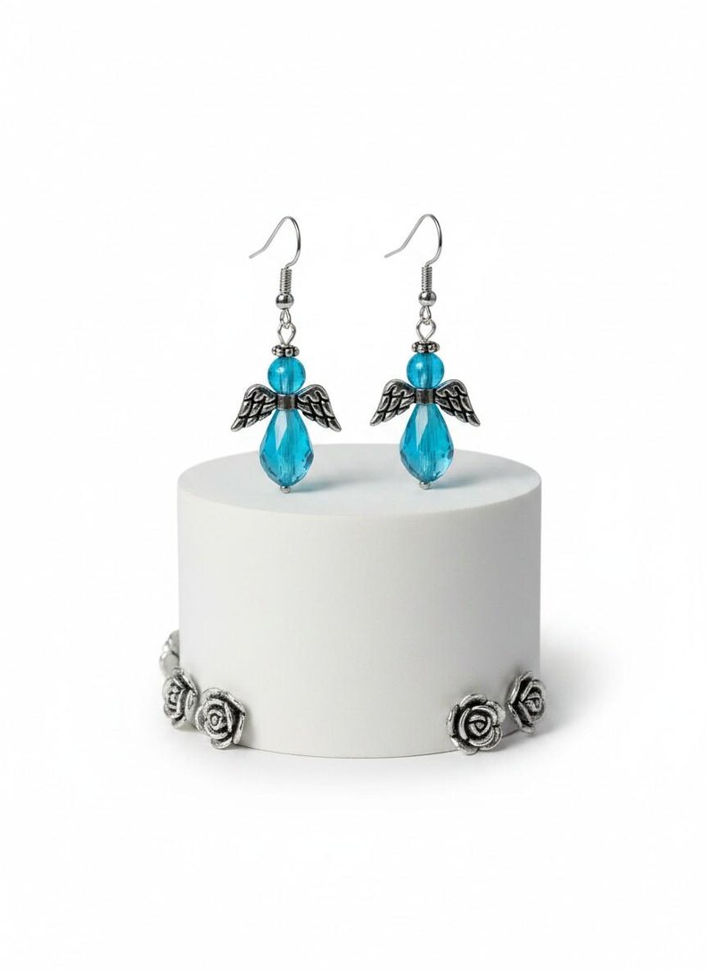 May include: A pair of angel-themed earrings with blue crystal teardrop pendants and silver-tone wings. The earrings are displayed on a white cylindrical stand with silver rose accents. The earrings have a hook closure.