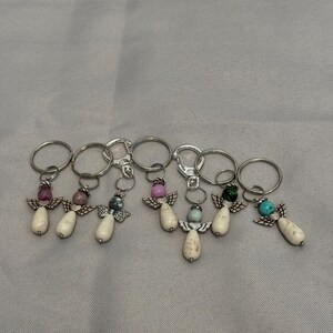 Puede incluir: Una colección de llaveros con forma de ángel. Cada llavero presenta un anillo plateado, un pequeño ángel con alas de cuentas y una cabeza de piedra preciosa. Las piedras preciosas son de varios colores, incluyendo rosa, azul y verde.