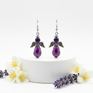 May include: A pair of angel-shaped earrings. Each earring features a purple teardrop-shaped bead, a smaller purple bead, and silver-colored metal wings. The earrings are displayed on a white pedestal, with white flowers and lavender sprigs in the foreground.