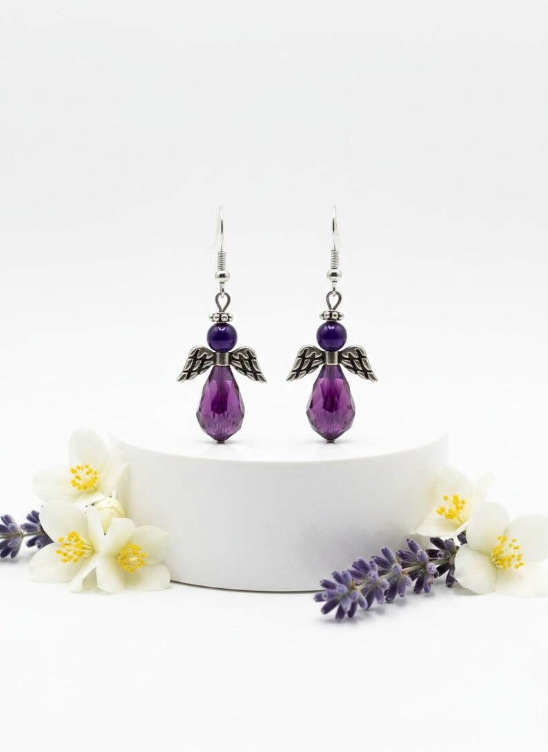 May include: A pair of angel-shaped earrings. Each earring features a purple teardrop-shaped bead, a smaller purple bead, and silver-colored metal wings. The earrings are displayed on a white pedestal, with white flowers and lavender sprigs in the foreground.