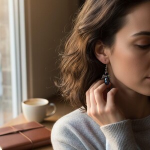 May include: A close-up of a dark blue crystal angel earring with silver-colored wings. The earring is dangling from a person's ear. The person is wearing a gray sweater. A cup of coffee and a brown leather notebook are on a wooden surface.