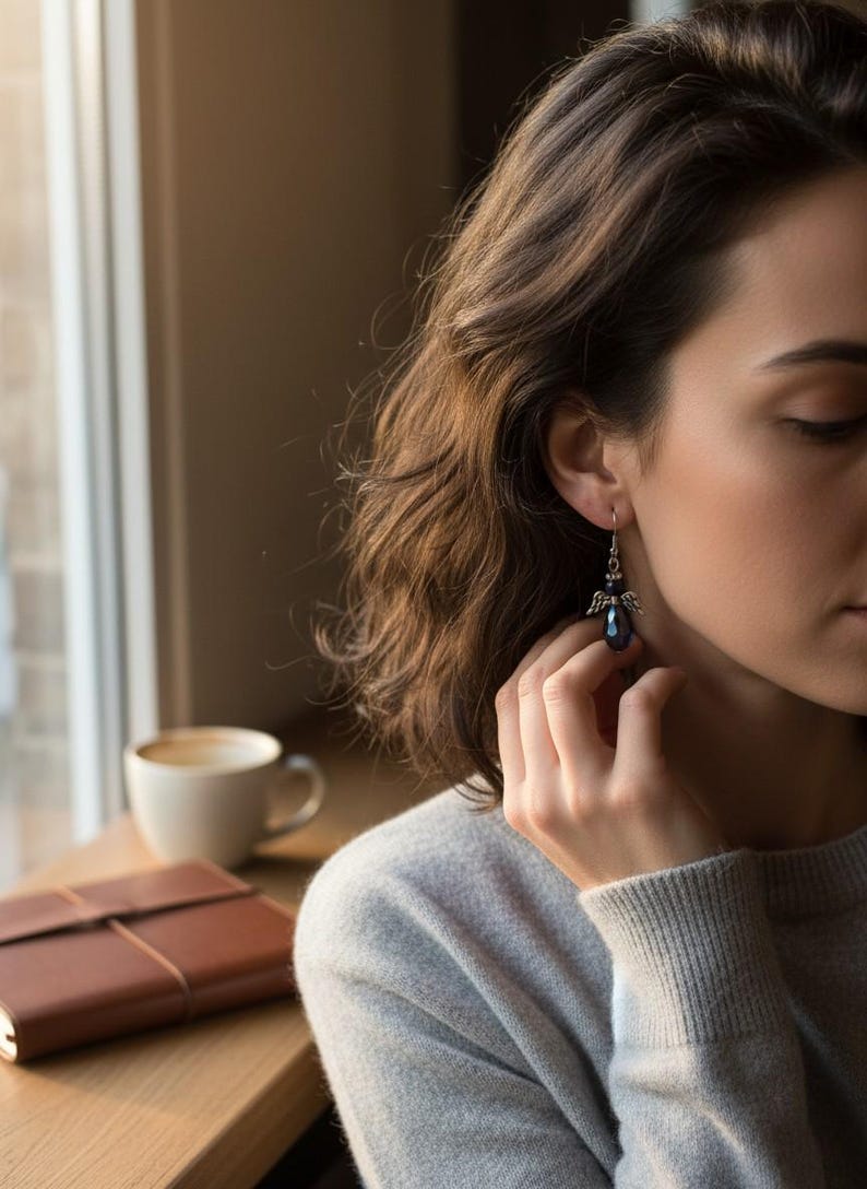 May include: A close-up of a dark blue crystal angel earring with silver-colored wings. The earring is dangling from a person's ear. The person is wearing a gray sweater. A cup of coffee and a brown leather notebook are on a wooden surface.