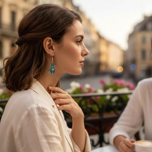 May include: A woman wearing a cream-colored blazer and silver earrings with blue crystal pendants. The earrings have a wing design above the crystal. The background shows a European city scene with buildings and flowers.