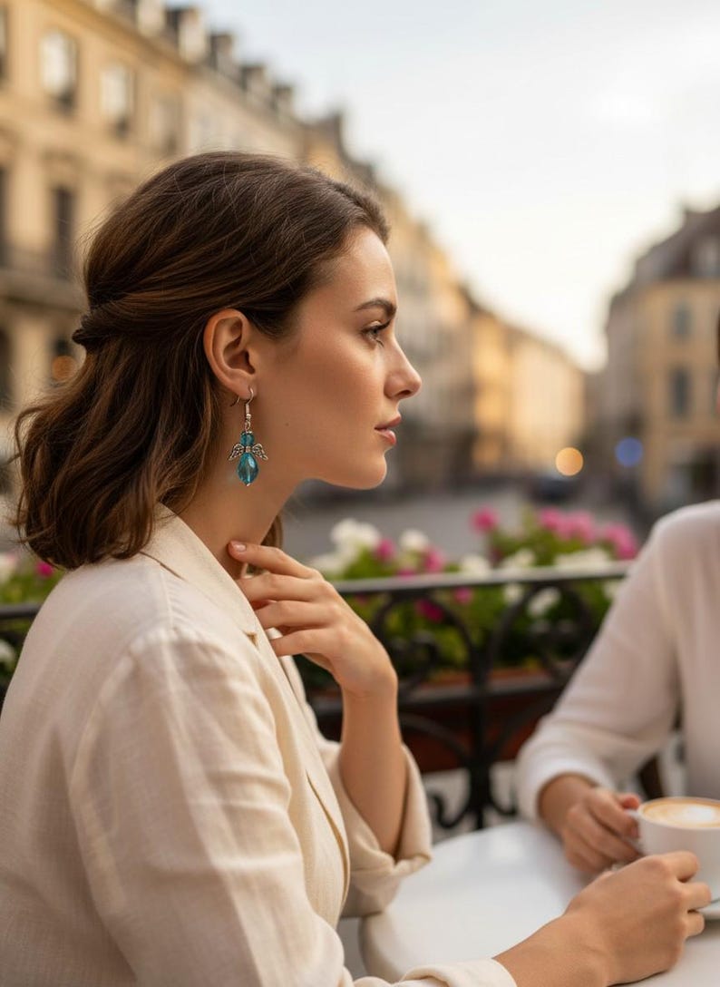 May include: A woman wearing a cream-colored blazer and silver earrings with blue crystal pendants. The earrings have a wing design above the crystal. The background shows a European city scene with buildings and flowers.