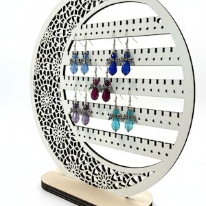 May include: A white wooden earring display stand with a decorative crescent moon shape. The stand holds several pairs of angel-themed earrings in shades of blue, purple, and teal. The stand has a wooden base and a detailed cutout design.