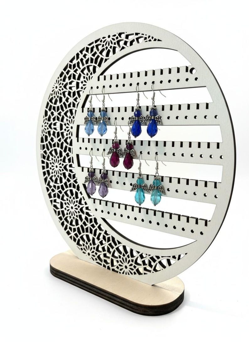 May include: A white wooden earring display stand with a decorative crescent moon shape. The stand holds several pairs of angel-themed earrings in shades of blue, purple, and teal. The stand has a wooden base and a detailed cutout design.