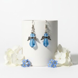 May include: A pair of angel-shaped earrings. Each earring features a blue, teardrop-shaped bead as the body, a smaller blue bead for the head, and silver-colored metal wings. The earrings are displayed on a white cylindrical object, with white and blue flowers in the foreground.