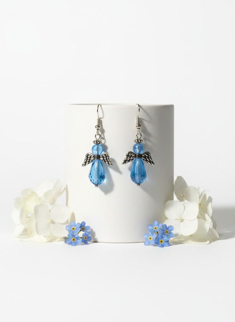 May include: A pair of angel-shaped earrings. Each earring features a blue, teardrop-shaped bead as the body, a smaller blue bead for the head, and silver-colored metal wings. The earrings are displayed on a white cylindrical object, with white and blue flowers in the foreground.