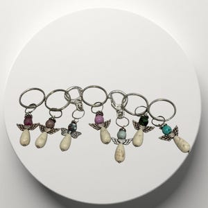 May include: A collection of angel-shaped keychains. Each keychain features a silver-colored ring, a teardrop-shaped bead, and small wings. The beads are in various colors, including pink, purple, blue, and green.