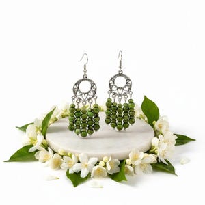 May include: A pair of silver-toned dangle earrings with green beaded accents. Each earring features a circular design with a floral pattern and multiple strands of small, round green beads. The earrings are displayed on a white marble surface, surrounded by white flowers.
