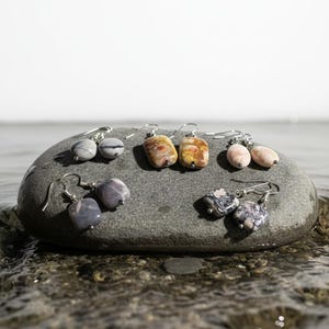 May include: Six pairs of gemstone earrings with silver hooks are displayed on a gray stone. The earrings feature various stone shapes and colors, including gray, brown, pink, and patterned designs. The stone rests in shallow water.
