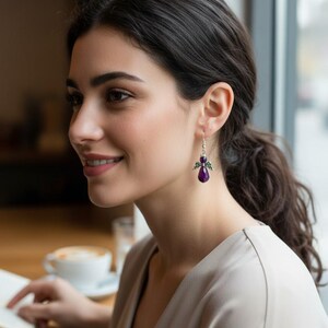 May include: A woman wearing a pair of silver dangle earrings with a purple gemstone and wing design. The earrings are the focal point, with the woman's profile and a blurred background of a cafe setting.