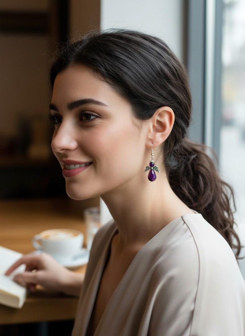May include: A woman wearing a pair of silver dangle earrings with a purple gemstone and wing design. The earrings are the focal point, with the woman's profile and a blurred background of a cafe setting.