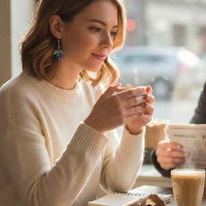 May include: A woman wearing a cream-colored sweater and blue angel earrings, holding a cup of coffee. A glass of coffee and a small bag of coffee beans are on the wooden table. The scene is set in a cafe with natural light.