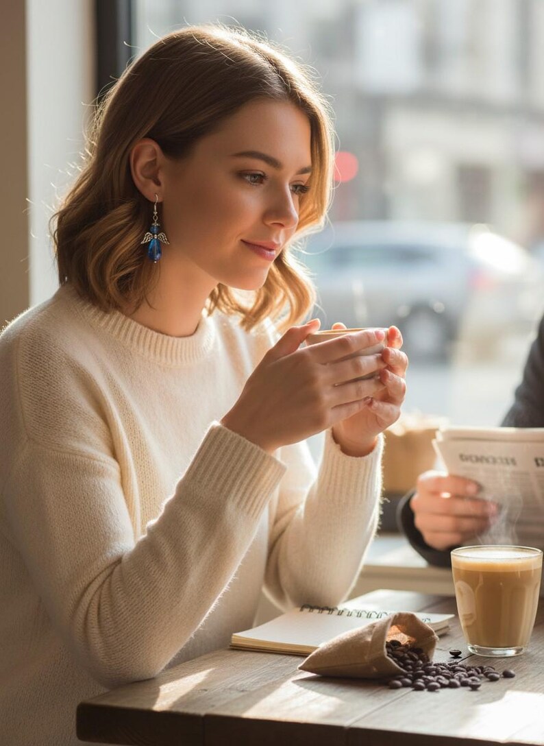 May include: A woman wearing a cream-colored sweater and blue angel earrings, holding a cup of coffee. A glass of coffee and a small bag of coffee beans are on the wooden table. The scene is set in a cafe with natural light.