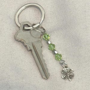 May include: A silver key with a decorative keychain. The keychain has green beads and a silver four-leaf clover charm. The key and keychain are on a light grey textured surface.