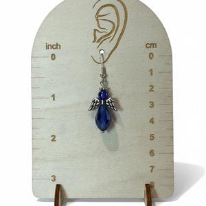 May include: A single blue angel earring with silver-colored wings and a faceted bead, displayed on a wooden earring display. The earring is approximately 1.5 inches long. The display has inch and cm markings.
