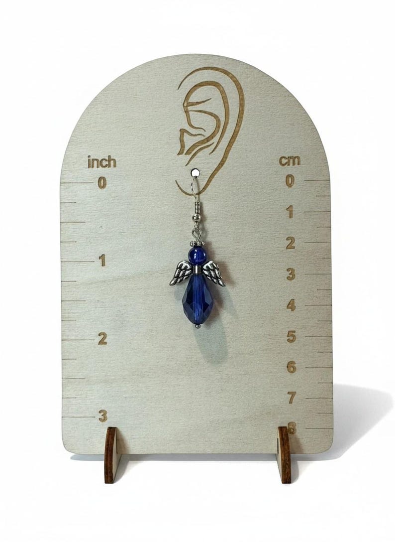 May include: A single blue angel earring with silver-colored wings and a faceted bead, displayed on a wooden earring display. The earring is approximately 1.5 inches long. The display has inch and cm markings.